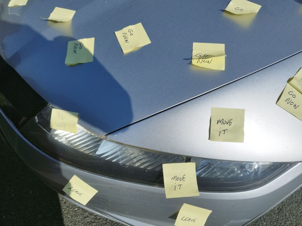 A close up of a car bonnet, upon which are stuck many post-it notes, reading things like: "move it", "go now", and "leave"