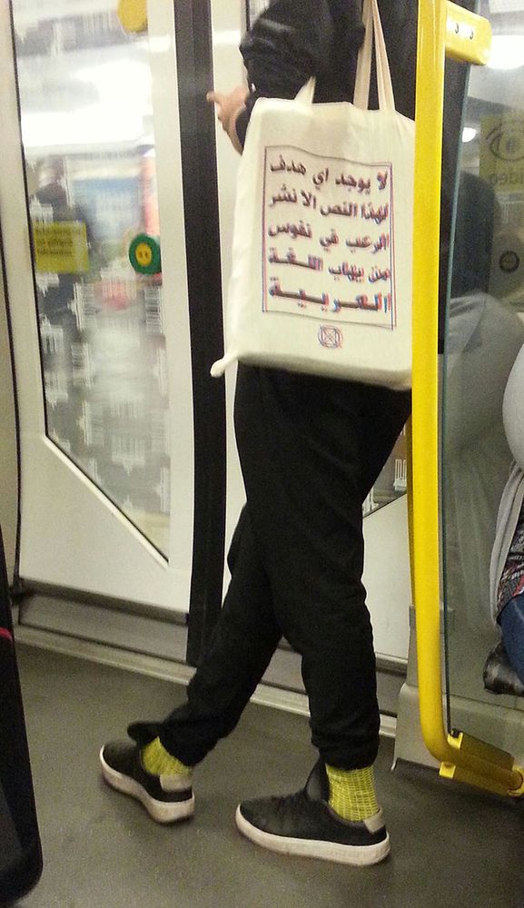 Photo inside a subway carriage. A person with a cloth bag over their shoulder. The bad has a body of Arabic Text printed on it.