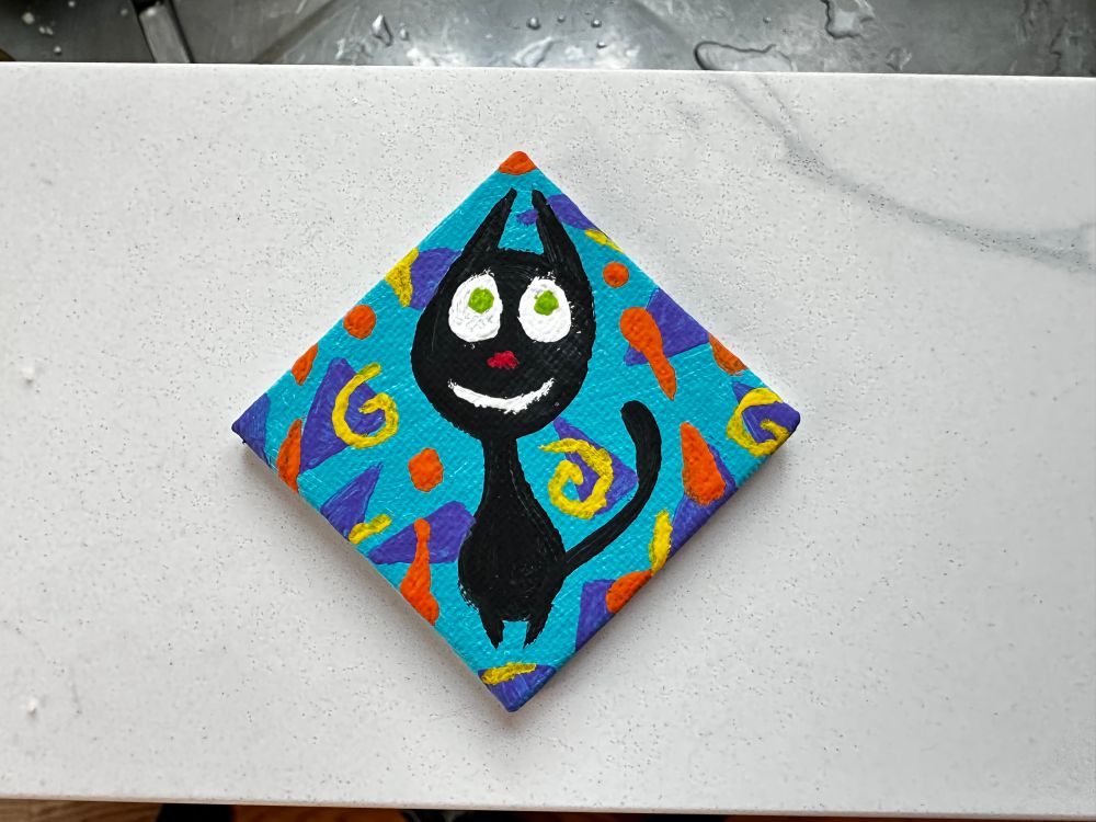 A little square canvas turned slightly so it’s diamond shaped. Painted is a teardrop shaped black cat with a red nose, big white eyes with light green pupils, and a turquoise background with purple triangles, orange drips, and yellow spirals