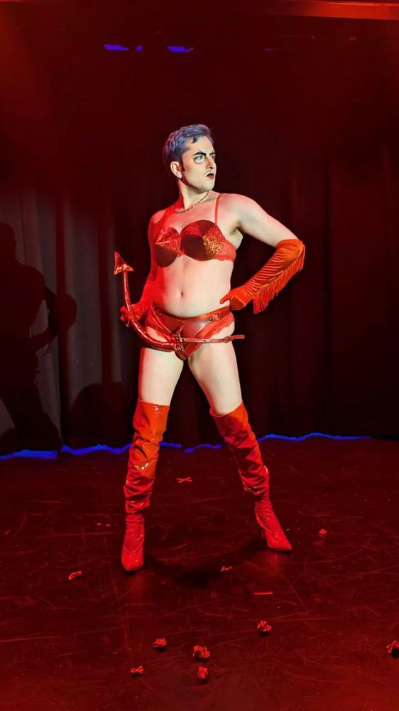 Drag king Zad Gravebone illuminated on the stage with dark blue short hair, sparkly, red bullet bra, red fringe gloves, a devil tail strap on, red thigh, high boots