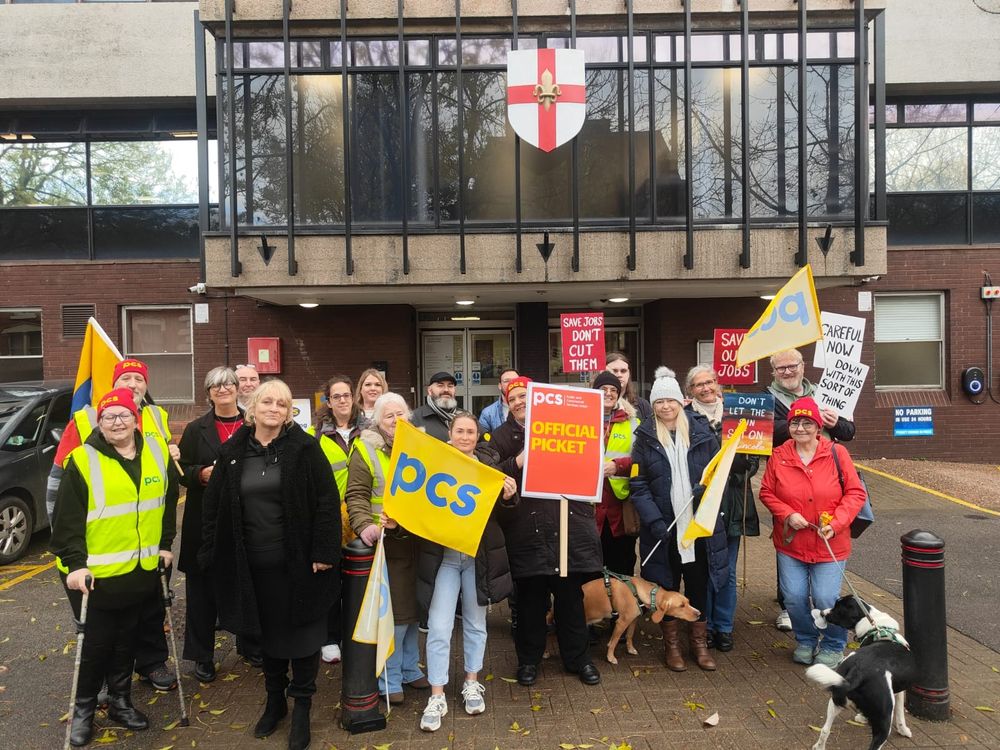 The photo shows 18 people and two dogs stood outside DWP Lincoln carrying placards and PCS flags, some wearing red PCS beanie hats.
