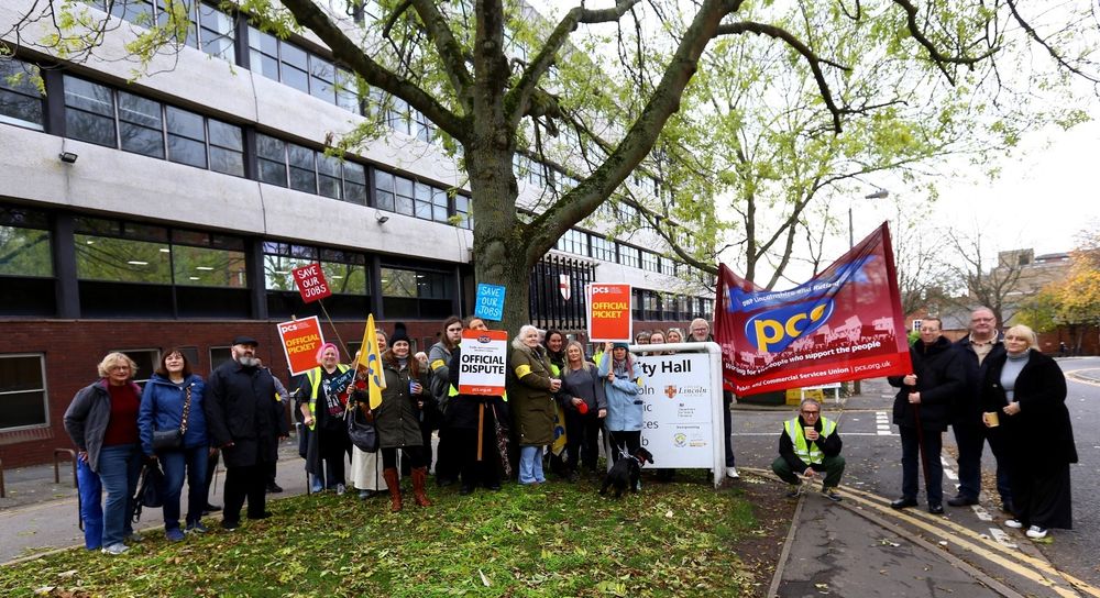 The photo shows around 20 people on a picket line outside DWP Lincoln holding a PCS banner and various placards.
