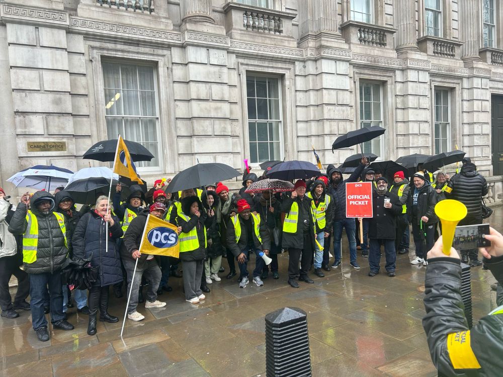 The photo shows around 25 people stood outside 70 Whitehall wearing high-vis jackets, holding PCS flags and umbrellas.