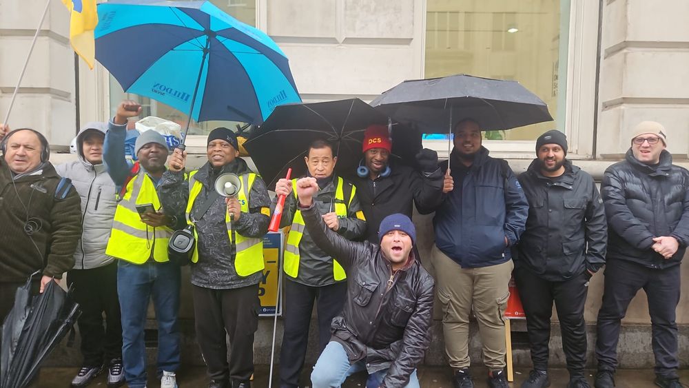 The photo shows 10 people on the picket line outside 22 Whitehall, 3 wearing high-vis jackets and 3 holding umbrellas.