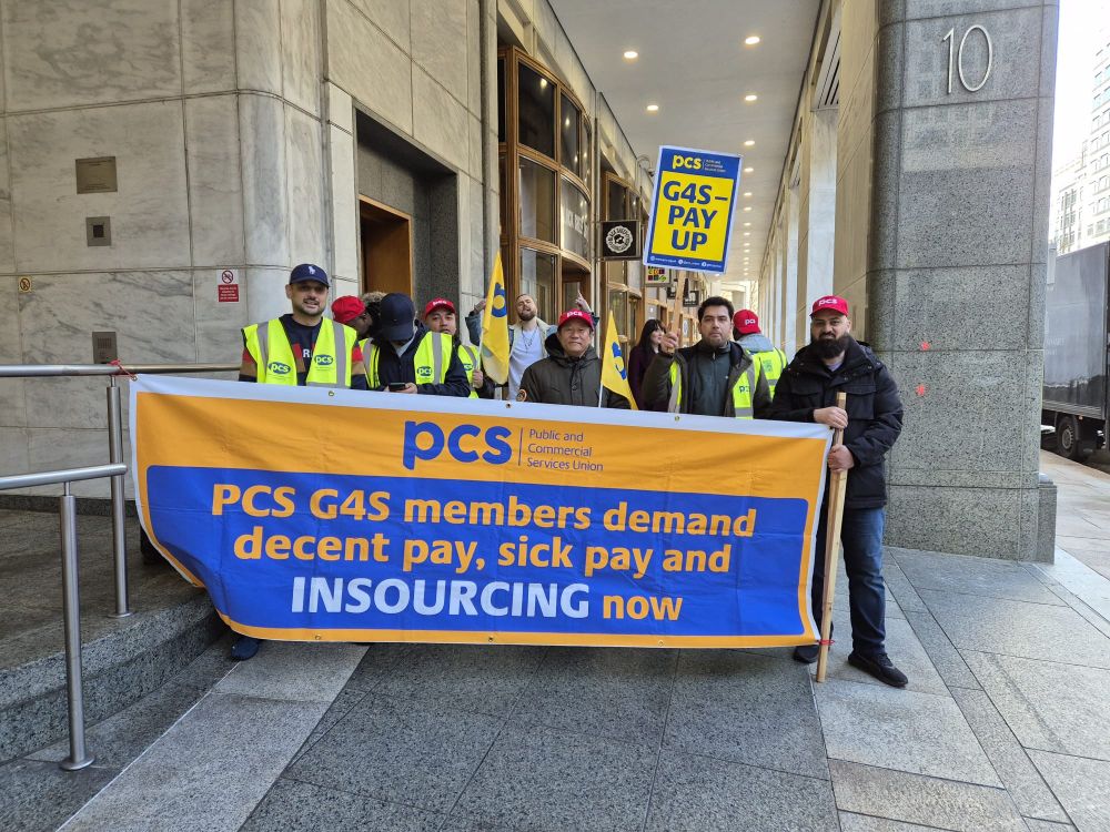The photo shows 11 strikers at 10 South Colonnade holding a banner which reads PCS G4S members demand decent pay, sick pay and insourcing now. They are also holding PCS flags, wearing high-rising vis jackets and one is holding a placard which reads G4S pay up. 