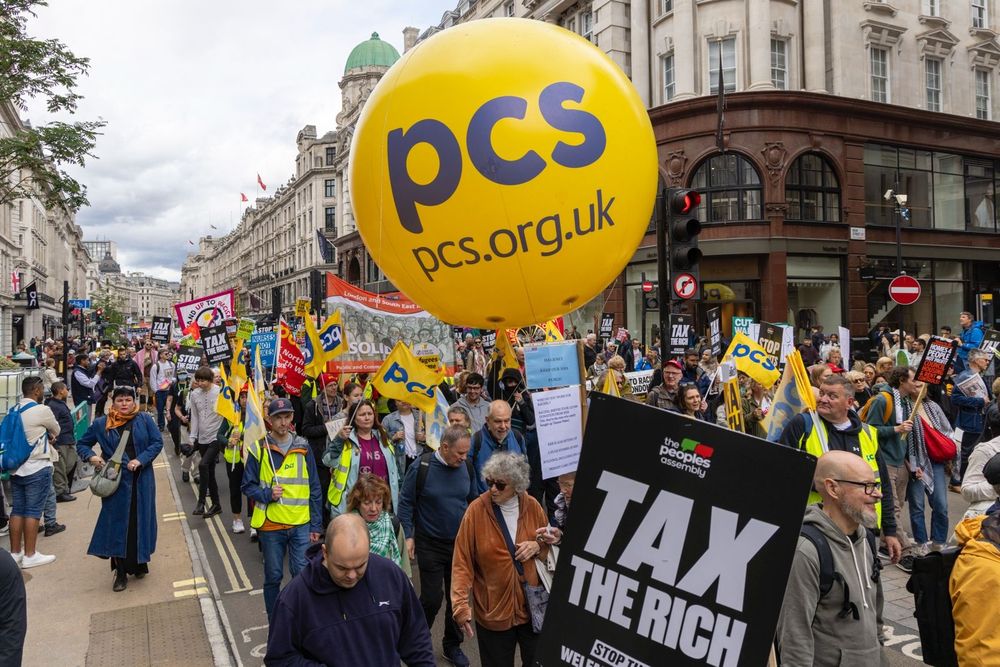 The photo shows a long line of marchers on the People's Assembly march in London with the PCS balloon in the foreground, alongside a People's Assembly banner which reads tax the rich