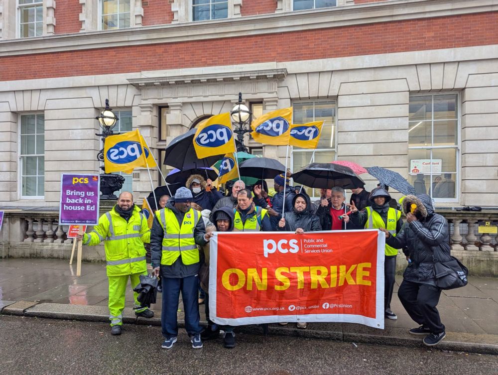 The photo shows around 20 people stood outside the Old Admiralty buildings holding a PCS on strike banner and wearing high-vis jackets and holding PCS flags and umbrellas.