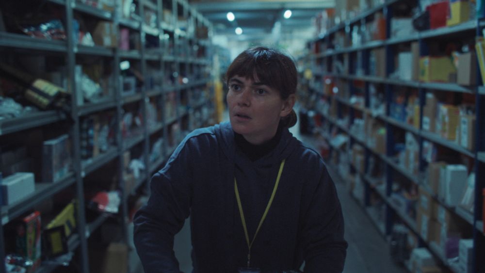 A still from the film showing the character Aurora in a dark warehouse with shelves stretching out on either side.