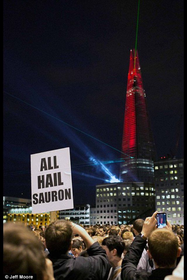 Above a number of grey London office buildings with white lighted windows, The Shard stands against the black night sky. Looming like a tapered spike, the tower is illuminated blood red. Spotlights and lasers shine outward from its base over the crowd, and a single green laser points upwards from its peak. In the foreground one member of the crowd has greeted the sinister spectacle with an upheld sign reading "All Hail Sauron".