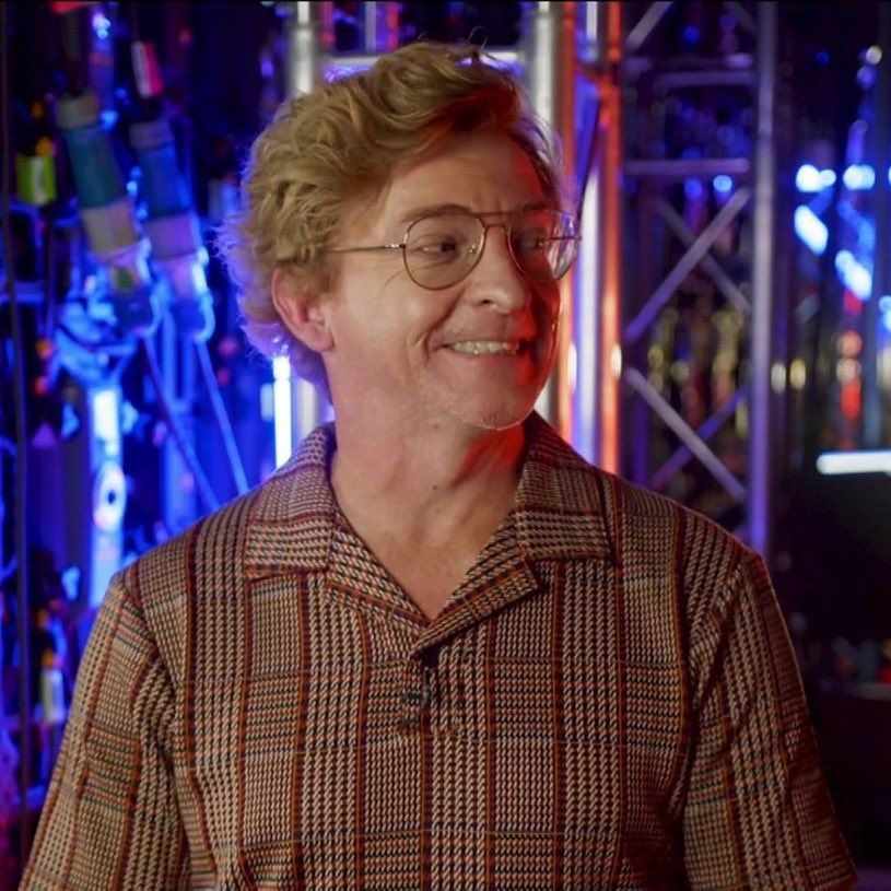 Picture or Rhys Darby with the sweetest smile, looking to his left. His hair is all fluffy, he has gold wire glasses and a brown, bronze and cream patterned polo that seems like a mix between houndstooth and tartan pattern.

The background is backstage material lit by blue lights. Rhys is lit by a neutral light from his right and a little bit of red light from the left.