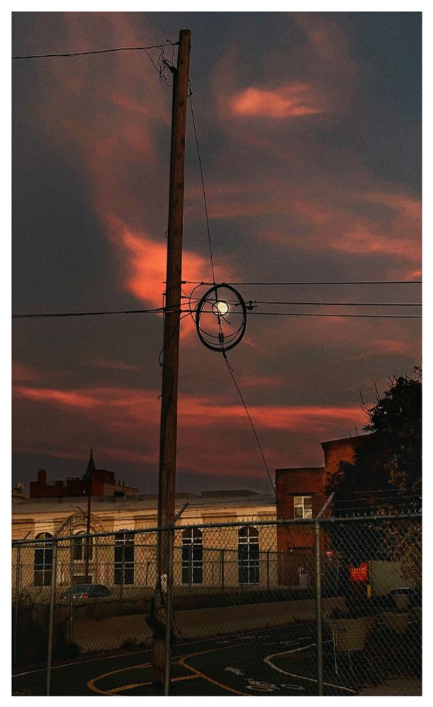 Photo of a city scene at sunset. Behind a chain link fence in the foreground, a road with yellow and white markings curves past some low buildings illuminated by the fading light. A tall wooden power pole rises into a darkening sky with luminous orange clouds. At the center of the image is the full moon, small in the sky, seen through some circular loops of cable attached to the power lines extending from the pole.