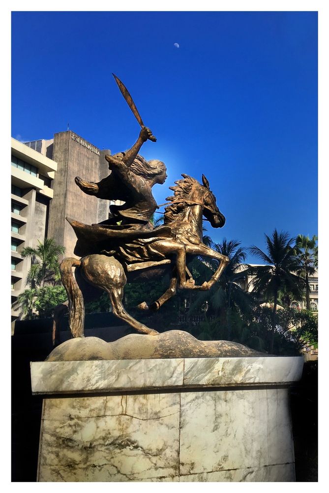 Photo of a bronze statue of a young woman, facing right and riding a horse that rears up with its forelegs raised. The woman's hair flows back as if wind-blown and she wields a raised bolo in her right hand. The statue stands on a wide marbled pedestal. Behind it is a line of palm trees and a high-rise building with the words THE PENINSULA at the top of its facade. High overhead in a blue sky, a tiny moon looks down on the scene.