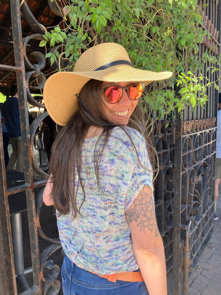 A woman poses in the Outline Crop Tee by Jessie Maed in front of a wrought iron fence covered in vinery. She is wearing sunglasses and floppy hat, looking back over her shoulder. The shirt is knit in a lace weight hand painted Manos del Uruguay yarn with pastel shades of blues, greens, purples and pinks. 