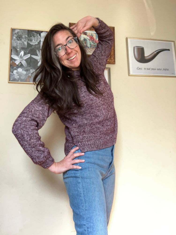 A woman poses excitedly in a newly finished hand knit sweater. The heathered purple yarn is somewhat mismatched; it was made before she learned about alternating skeins or how to purl correctly, but she is full of pride anyways for finishing her first handmade garment. 