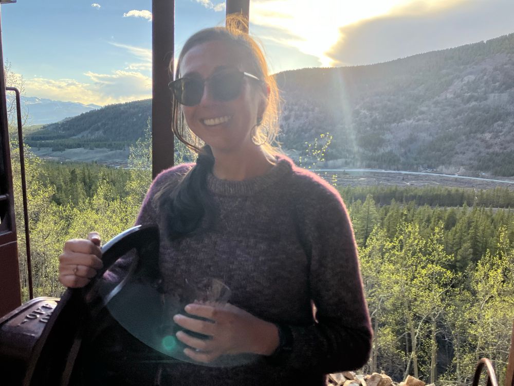 A woman poses in front of the setting sun with a mountain valley backdrop. The saturated light makes the aspen leaves sparkle. She is wearing a hand knit purple sweater that was finished that morning, ends woven in on the car ride. 