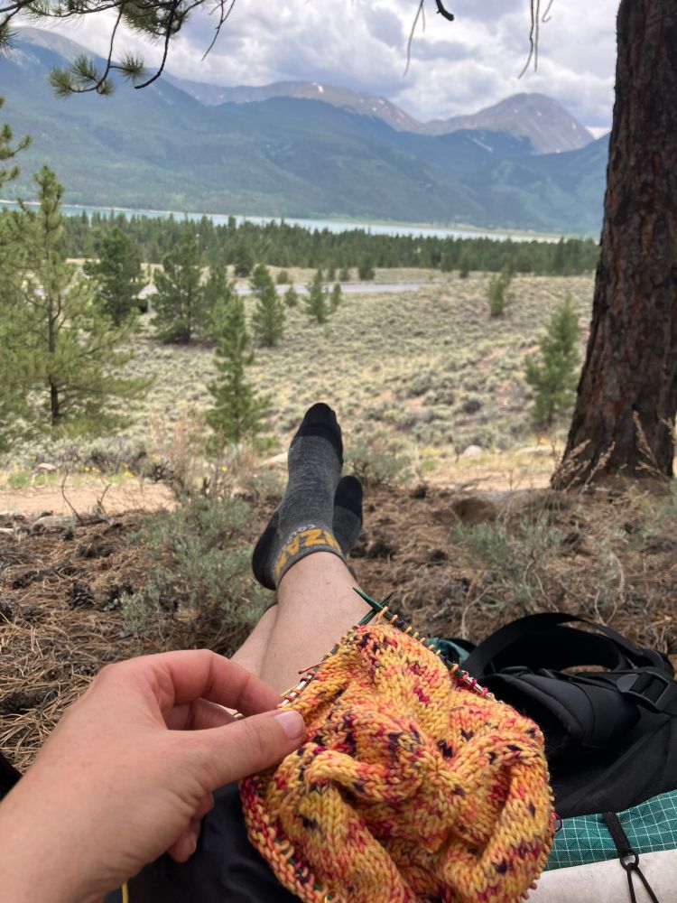 The beginnings of a Musselburgh hat, pattern by Ysolda, on small needles in a lap. Legs are propped up on a backpack, hiking shoes are off for a break under a tree in a small ponderosa forest. In the background is a lake and many mountains, including the final pass of the loop trail: a beastly climb to finish out the last day of a long long walk, still over a week away. 