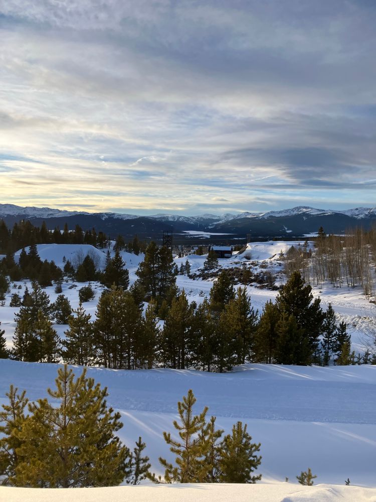 Dramatic clouds hover over a broad mountain landscape. A frozen lake is visible in the distance. The ground is covered in undisturbed snow and pine trees cluster throughout. 