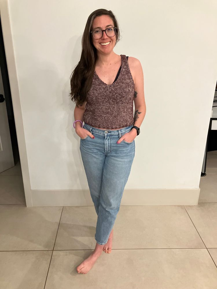 A woman in glasses and blue jeans stands in front of a white wall modeling a Wisp tank top by Caitlin Hunter. The straps were modified in the back to be more of a racer back style. The tank top is knit with a heathered purple cotton yarn. 