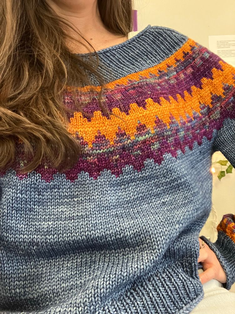 My Easy V by Caitlin Hunter also features the malabrigo sunset color way in a worsted weight: close up of a denim blue v neck sweater with zigzag color work in a deep golden yellow, a variegated purple and aqua, and purple stripes on the yoke and sleeve cuffs. 