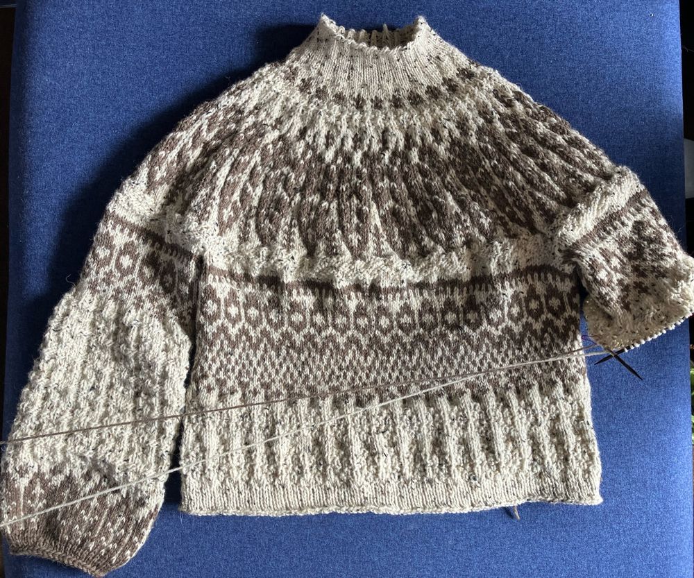 A hand knit sweater, in progress, on a blue background. The sweater is cream colored with brown patterns and varied texture all over. 
