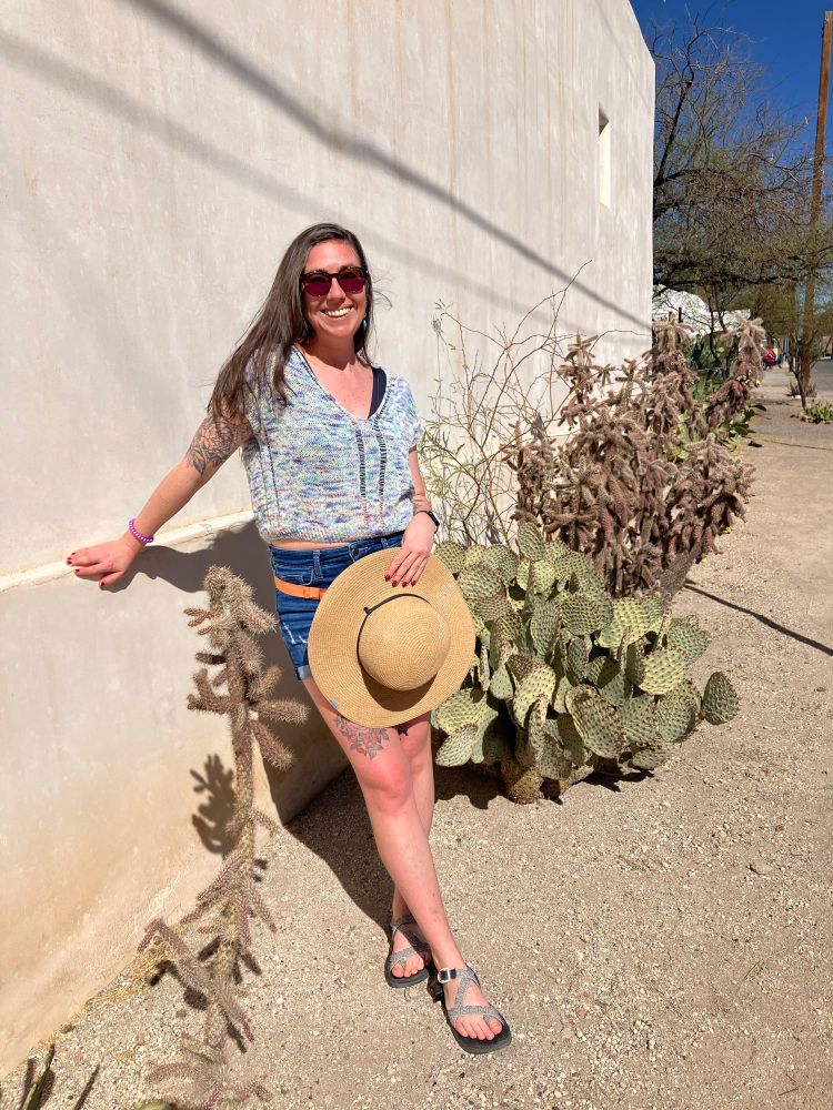 A woman leans against an abode wall in front of a variety of cacti, modeling a hand knit short sleeve shirt