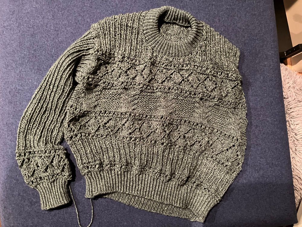 A nearly finished gray sweater lays on a blue ottoman. 

I left quite a bit of yarn on the right sleeve and didn’t bind off, then did the neck finishing before moving on to the left sleeve. I wanted to make sure the neck didn’t pull up the sleeves and make them too short- I love a sweater I can pull over my hands! This turned out perfectly cozy. 