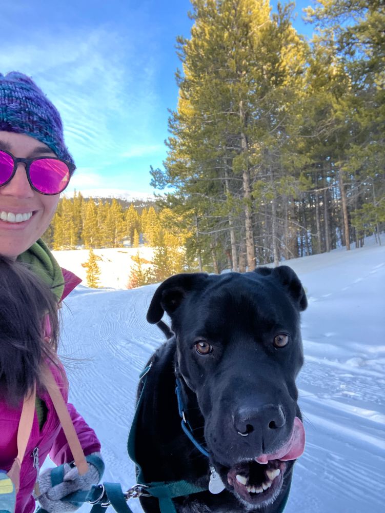 A woman in sunglasses tries to get her usually photogenic black dog to pose for a selfie. He is in motion, moving towards the camera with his tongue out sideways. 