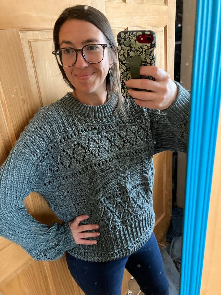 A woman poses proudly in a finished hand knit gray sweater with eyelets and cables. This is the Salty Days pattern by Veronika Lindberg, knit with Lion brand Jeans yarn. 