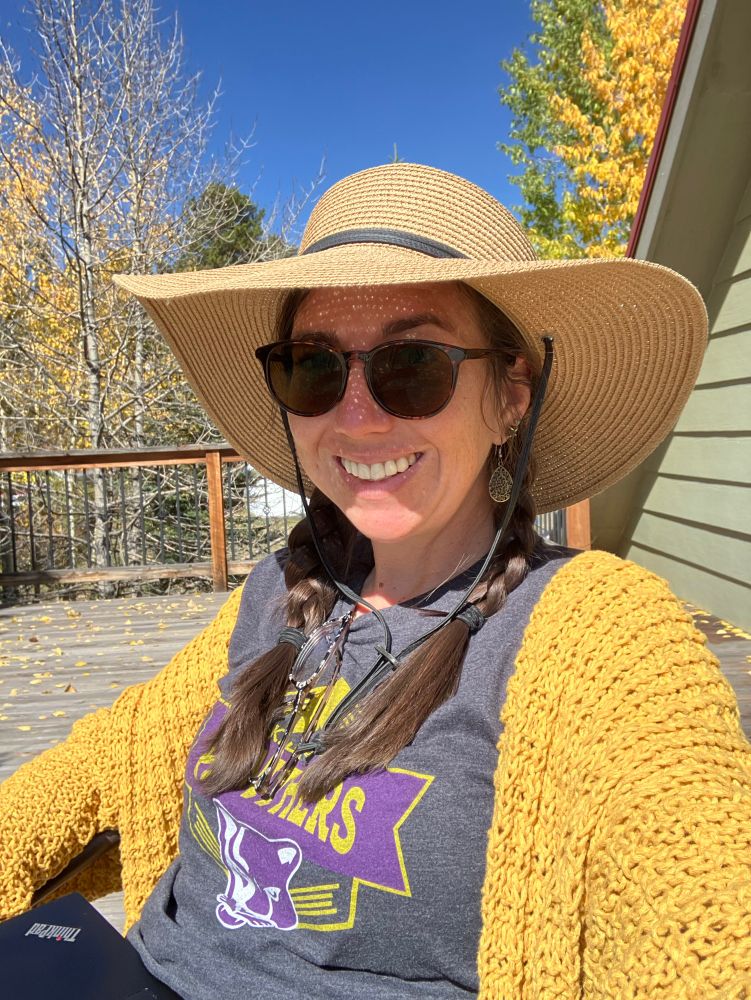 A woman in a yellow hand knit cardigan, large hat and sunglasses smiles on a wood deck. In the background, a variety of aspen and cottonwood are visible. The yellow fall leaves match the color of the cardigan. 