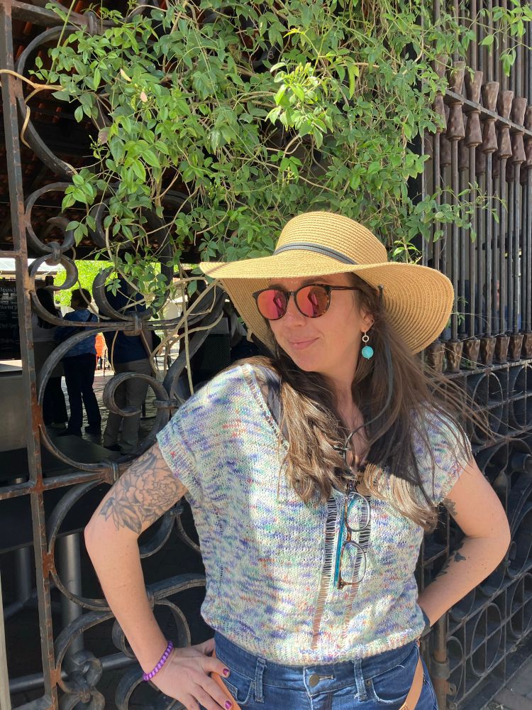 A woman in a large floppy hat and sunglasses looks over her shoulder, smiling, in front of a wrought iron fence overgrown with leafy green vines. She is modeling a hand knit short sleeve shirt made with lace weight painted yarn featuring a variety of pastel hued colors. 