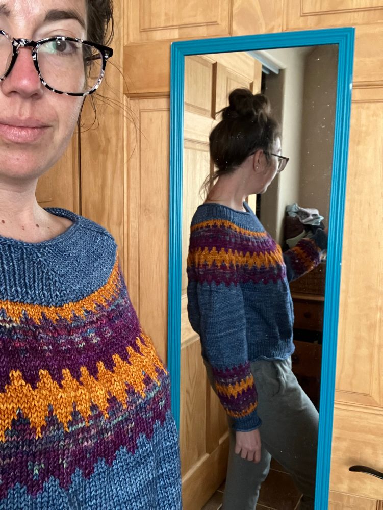 A woman tries on a newly finished Easy V sweater, fresh off the needles and before blocking. She stands in front of a mirror to see the side profile of the sweater, cropped slightly to hit at the hip. The sweater is a denim blue with zigzagging stripes of golden yellow, tonal purple/teal/pink, and rich plum on the yoke and cuffs. The hems of the sweater feature a roll past the ribbing giving the sweater a relaxed and flowing fit. 