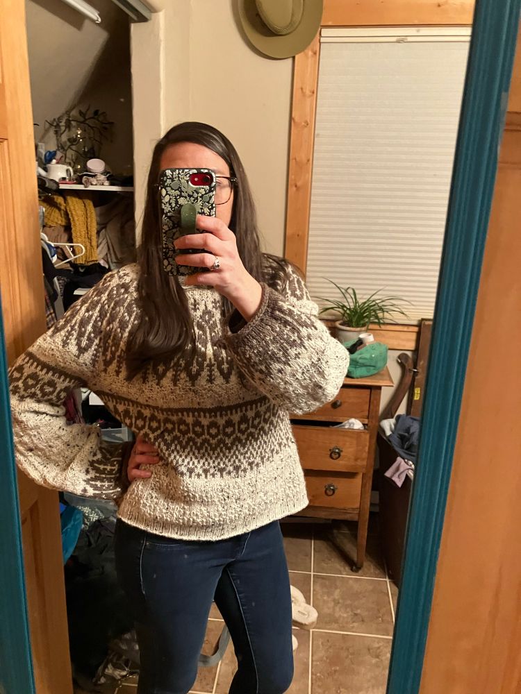 A woman poses in a hand knit long sleeve cream colored sweater with brown patterns all over. 