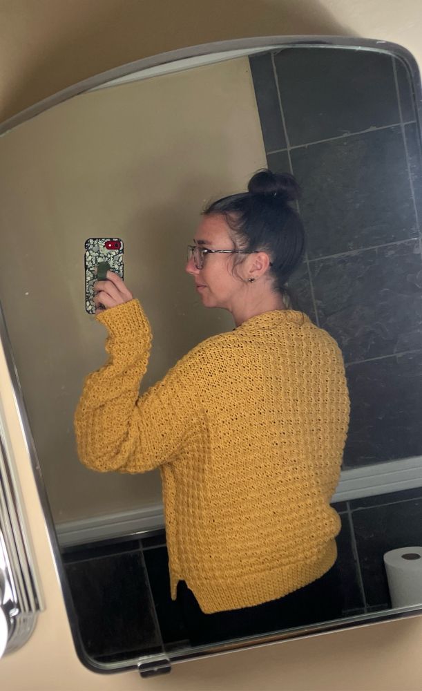 A woman in glasses models the back of a hand knit yellow cardigan in an all over waffle texture. The cardigan has a slightly oversized, boxy fit. 