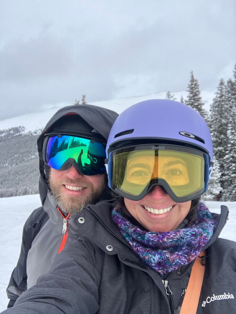 A man and a woman in ski goggles and helmets smile in the foreground of a snowy, stormy day on the slopes. Behind them, a snow covered mountain looms with pine forest up to the tree line. 