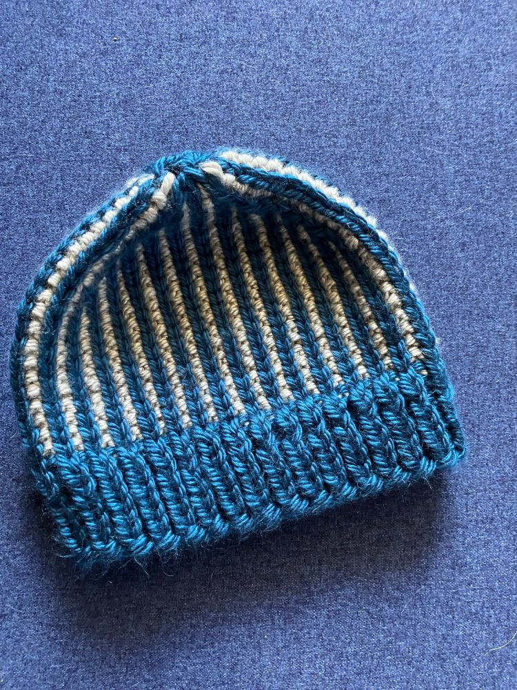 A beanie with vertical blue and gray stripes and a blue brim lays on a darker blue background. I made this version of the Humdrum hat for my FIL figure- the alternating color in the ribbing made for a much stiffer and less squishy hat than I aimed for, so he may be receiving a second attempt this year! 