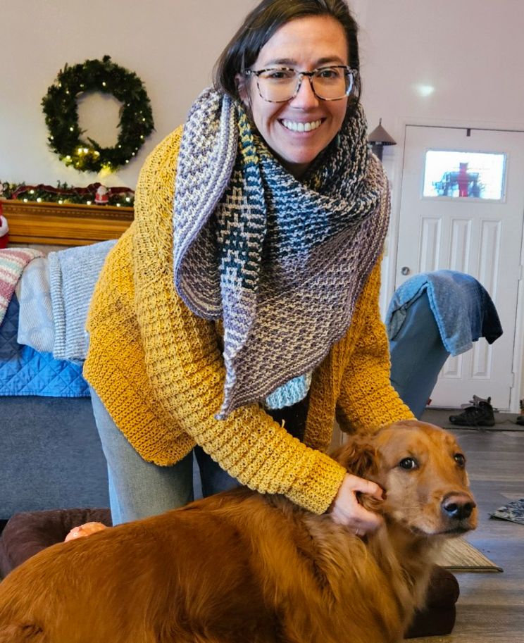 A woman plays with a dog, she is wearing a yellow hand knit cardigan and a mosaic shawl designed by Audrey Borrego in shades of purple, grey, white, teal, and yellow. 