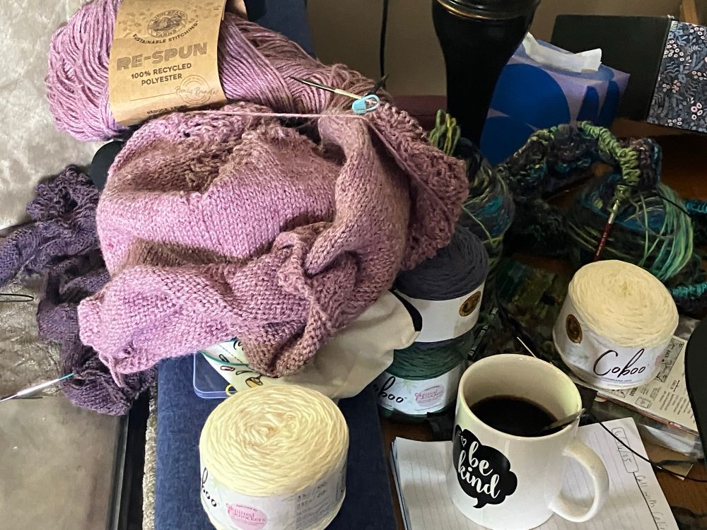 A crowded couch arm and coffee table are pictured, cluttered with a variety of knitting projects, coffee, maps, and backpacking guides. On top of the pile is the Provence Top in a dusty pink. Beneath that is the beginning of a Lenore cardigan by Audrey Borrego in a rich heathered purple. In the forefront are a few skeins of cotton bamboo blend destined for a Zinnie Tee, also by Audrey Borrego, in white with indigo stripes and green squares. Bear the back of the table by a lamp is a large ball of variegated yarn destined to be a dog blanket. This is a recently frogged project, done entirely in stockinette when I was a novice knitter, that I’ve pulled out to make a border of moss stitch and some purpled detail stripes throughout. A less typical WIP is the planning for a long walk I’ll do this summer with a friend, and perhaps I’ll bring some knitting along!