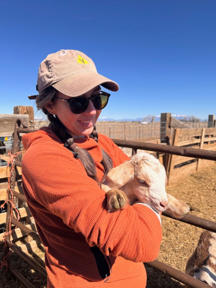 A woman in an old cap and sunglasses holds a baby goat in a fenced pen. Mountains loom in the distance 