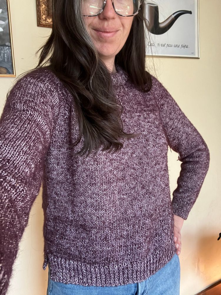 A woman poses in a newly finished hand knit sweater. The heathered purple yarn is somewhat mismatched; it was made before she learned about alternating skeins or how to purl correctly, but she is full of pride anyways for finishing her first handmade garment. 