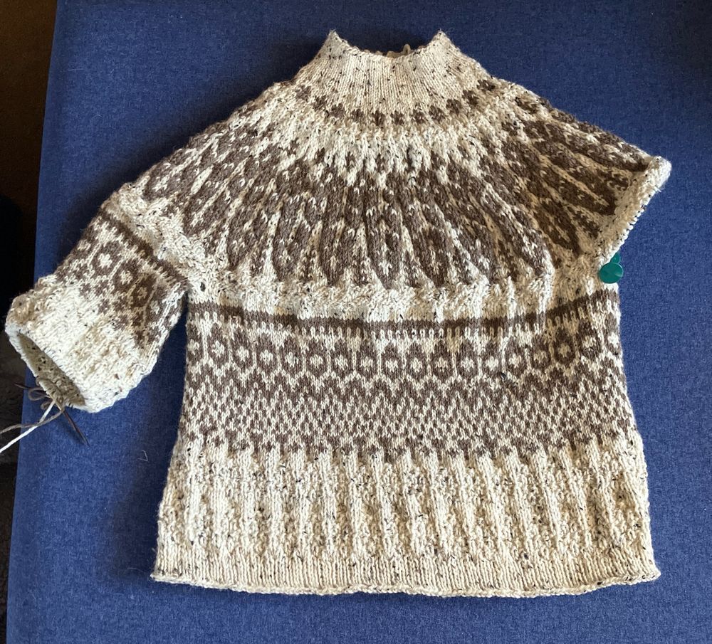 A hand knit cream colored sweater with brown patterns all over is nearly finished, needing one and a half sleeves. 