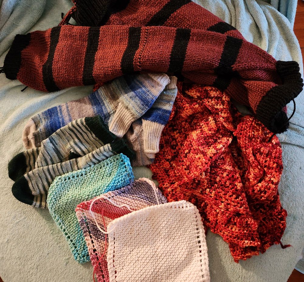 pile of almost finished knitting projects; red and black striped sweater, blue and grey socks, multi colored green socks, 3 dishcloths, and fire colored crochet shawl. 
