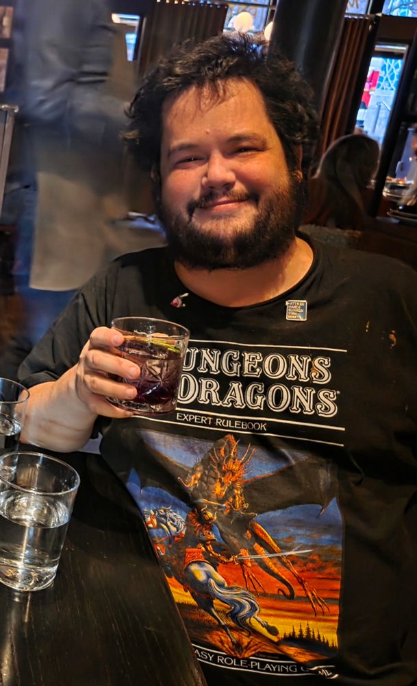 Me at this fancy tapas place drinking a fancy drink in my D&D expert handbook shirt with two pins: one is a Final Fantasy type menu pin and the other is a Kirby ate Link Smash pin