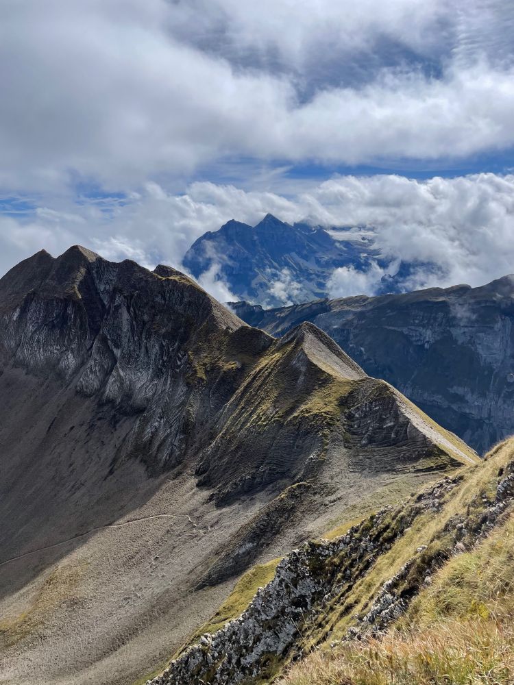 Blick von unterhalb eines Berggipfels in Richtung eines ausgesetzten Grats, im Hintergrund noch höhere Berge, vielfältige Wolken