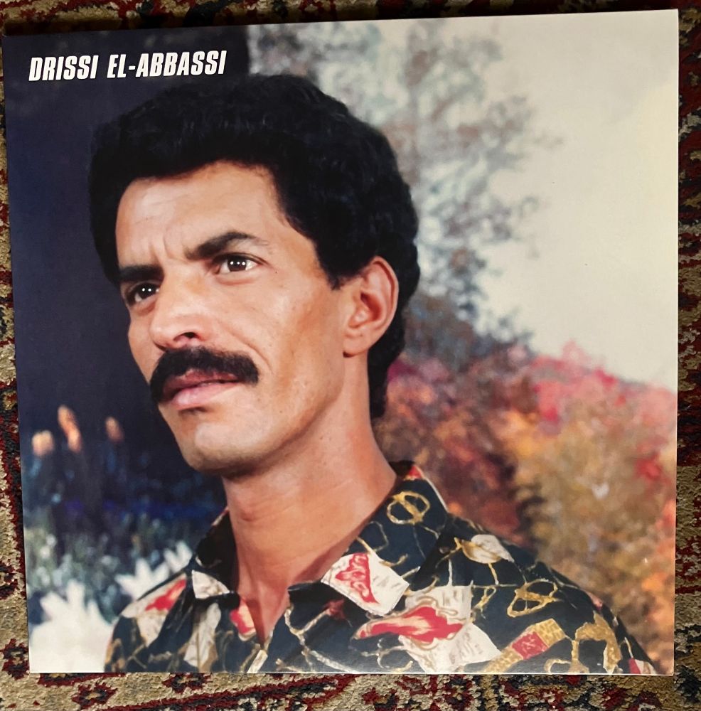 Head and shoulders colour photo of a moustachioed Drissi El-Anbassi resplendent in black/gold/red/white patterned shirt 