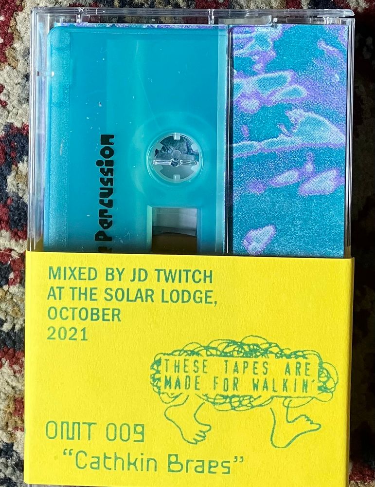 Back of cassette. Aqua Blue cassette. Thick yellow band around the bottom half. Blue/green text reads:
MIXED BY JD TWITCH AT THE SOLAR LODGE, OCTOBER 2021 
THESE TAPES ARE MADE FOR WALKIN’
OMT009
“Cathkin Braes”