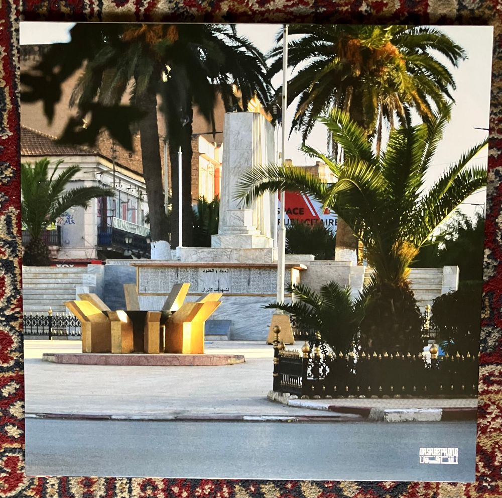 Colour photo of a street scene with palm trees, grey marble(?) monument and gold sculpture/table/seat. Tiled roof building in the background. 