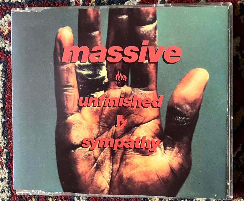 Front cover of CD.
A scarred, lined and weathered black hand, palm facing forward, bandaged fingers raised on a greyish, greenish, blueish background. 
Red text reads: 
massive 
unfinished 
sympathy 