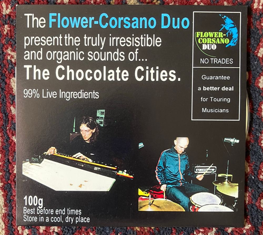 Black sleeve with white and blue text and 2 colour photos. One shows Mick Flower playing the Japan Banjo, the other shows Chris Corsano playing drums. 
Text reads: The Flower-Corsano Duo present the truly irresistible and organic sounds of...
The Chocolate Cities.
99% Live Ingredients
FLOWER-CORSANO DUO NO TRADES
Guarantee a better deal for Touring Musicians 
100g
Best before end times
Store in a cool,dry place