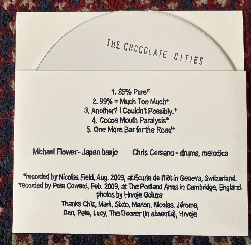 Back of CD sleeve.
White CDr with CHOCOLATE CITIES stamped in black text
Track listing in black text on folded white paper reads: 
   1.  85% Pure*
2. 99% = Much Too Much*
3.    Another? I Couldn't Possibly.*
4.    Cocoa Mouth Paralysis*
5.    One More Bar forthe Road*

Michael Flower - Japan banjo 
Chris Corsano - drums, melodica

*recorded by Nicolas Field, Aug. 2009, at Ecurie de I'llat in Geneva, Switzerland. recorded by Pete Coward, Feb. 2009, at The Portland Arms in Cambridge, England.
Pnotos by Hraje Golura
Thanks Chiz, Mark, Sixto, Marion, Nicolas, Jérome, Dan, Pete, Lucy, The Doozer (in absentia), Hivoje
