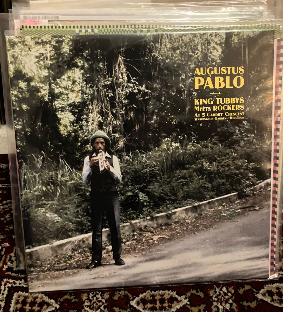 Augustus Pablo standing on a road in front of some jungle vegetation, playing a melodica 